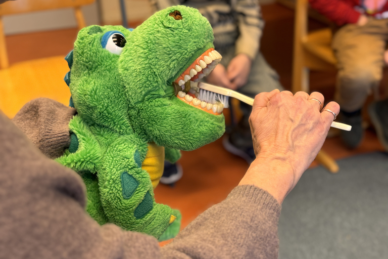 Es ist eine grüne Krokodilhandpuppe zu sehen. Eine Frau zeigt am Krokodil, wie man sich die Zähne richtig putzt